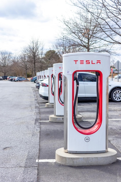 A row of Tesla Supercharger stations, each with a Tesla vehicle plugged in. The stations are clean and well-maintained, with clear signage. The sky is a vibrant blue, and there are trees in the background.