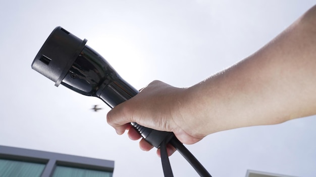 A close-up shot of a hand plugging an EV charger into an electric vehicle at a public charging station. The charging station screen displays the charging progress and available payment options. The focus is on the connection point and the charging cable.